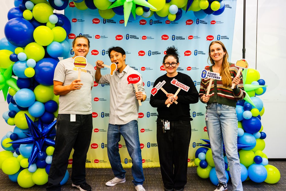 Volunteers holding photo props in front of the balloon backdrop at the Blessings in a Backpack meal-packing event