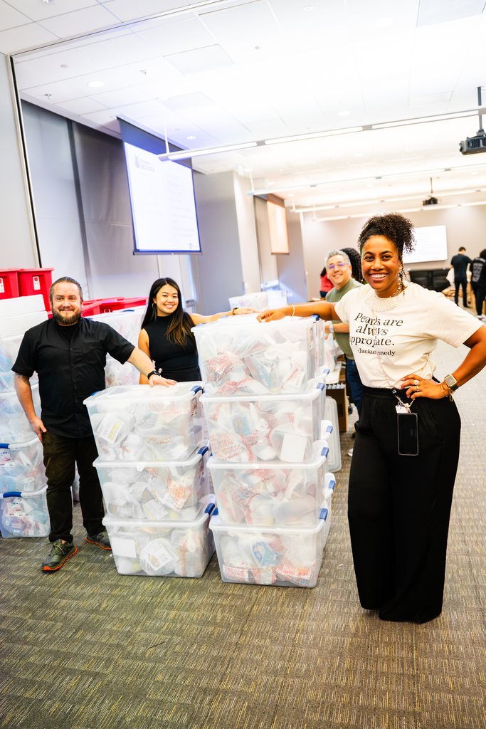 Volunteers standing beside bins of packed weekend meal bags for Blessings in a Backpack